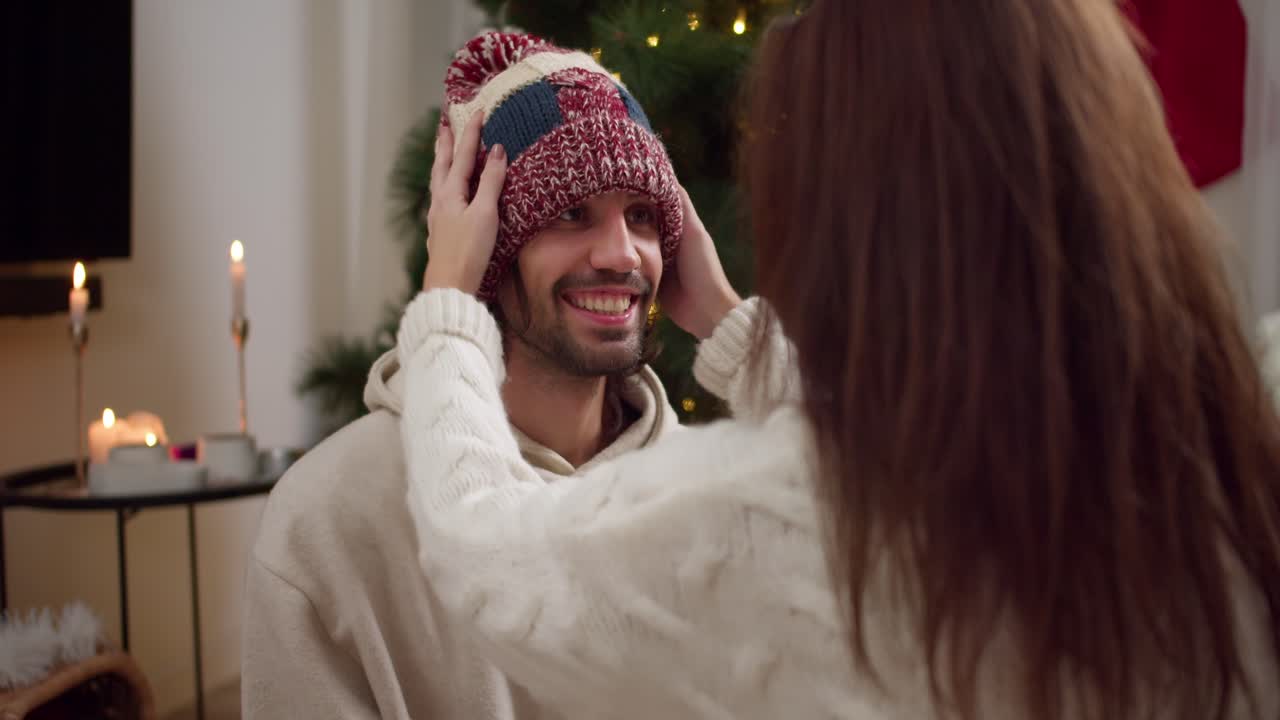 sobre el hombro: una chica morena con un suéter blanco pone un sombrero de lana roja en la cabeza de su novio cerca del árbol de año nuevo en una habitación acogedora decorada para el año nuevo