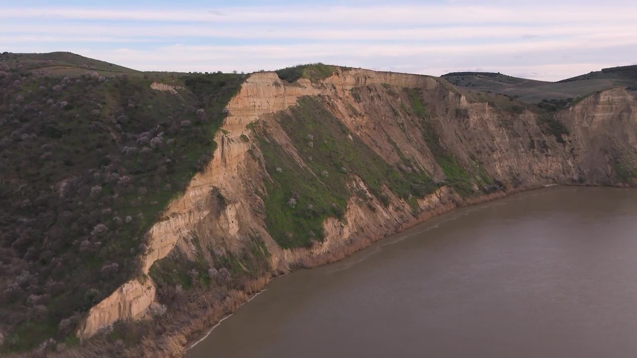 Ripresa aerea con drone che vola lateralmente e sale dal fiume Tago, concentrandosi su una ripida scogliera verticale con vegetazione verde, sopra acque torbide.