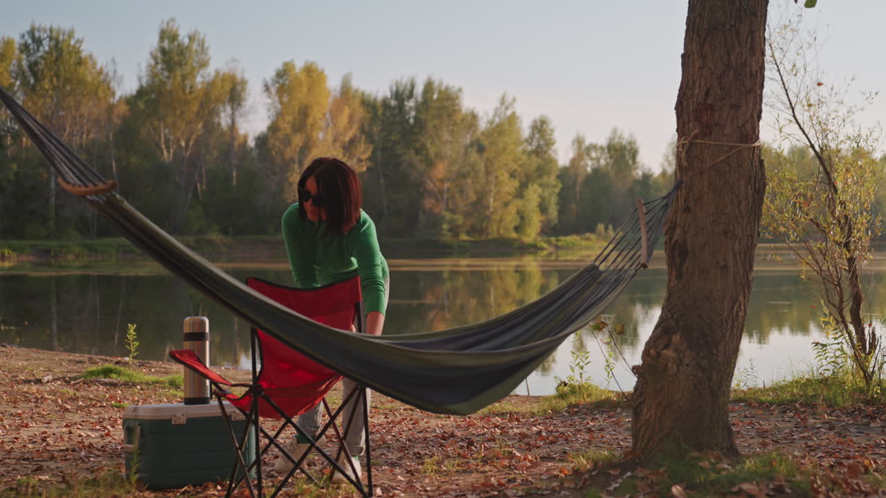 Donna bianca si rilassa su amaca da campeggio in riva al lago, sorseggia da una tazza, osserva l'acqua, brezza leggera tra gli alberi, borsa frigo e sedia accanto all'amaca, luce dorata del crepuscolo, solitudine tranquilla e lento distendersi all'aperto.