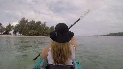 girl with long blonde hair wearing a black hat paddling a kayak near a tropical island