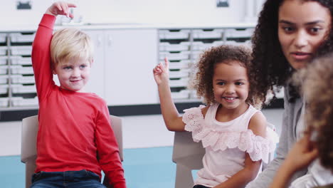 infant school children raising hands to speak in a lesson with their female teacher, close up