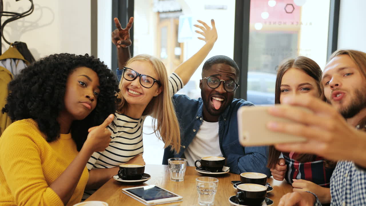 vista de cerca de un grupo multiétnico de amigos haciendo un selfie con un smartphone y haciendo gestos divertidos sentados en una mesa en un café