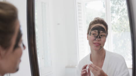 Happy-biracial-woman-applying-beauty-face-mask-looking-at-mirror-in-bathroom,-slow-motion