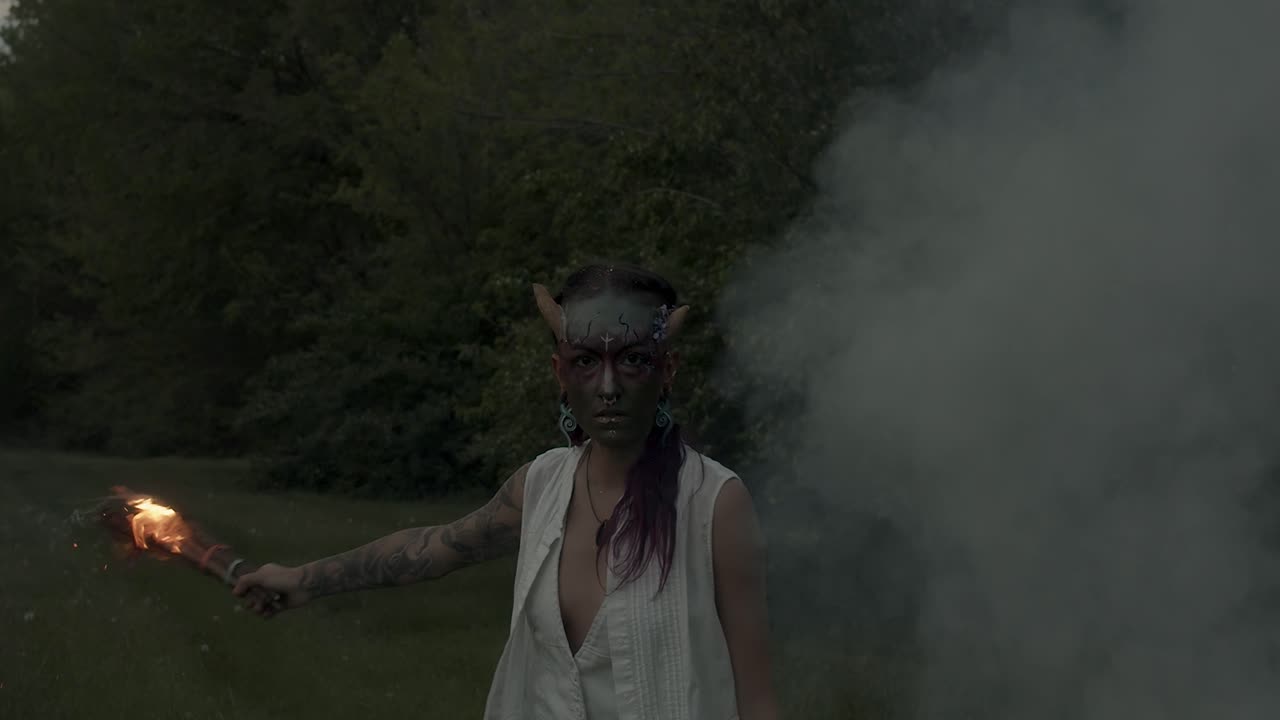 mujer con pintura facial de beltane empuja una antorcha de fuego cerca de una hoguera humeante en un festival pagano nocturno