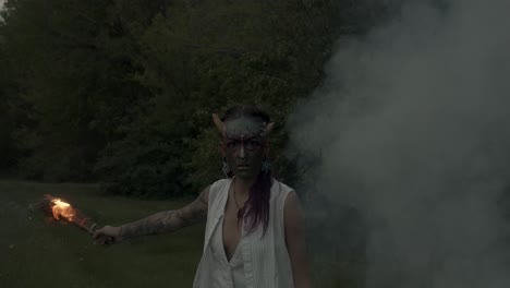 woman with beltane face-paint wields torch of fire near a smoking bonfire at a night pagan festival