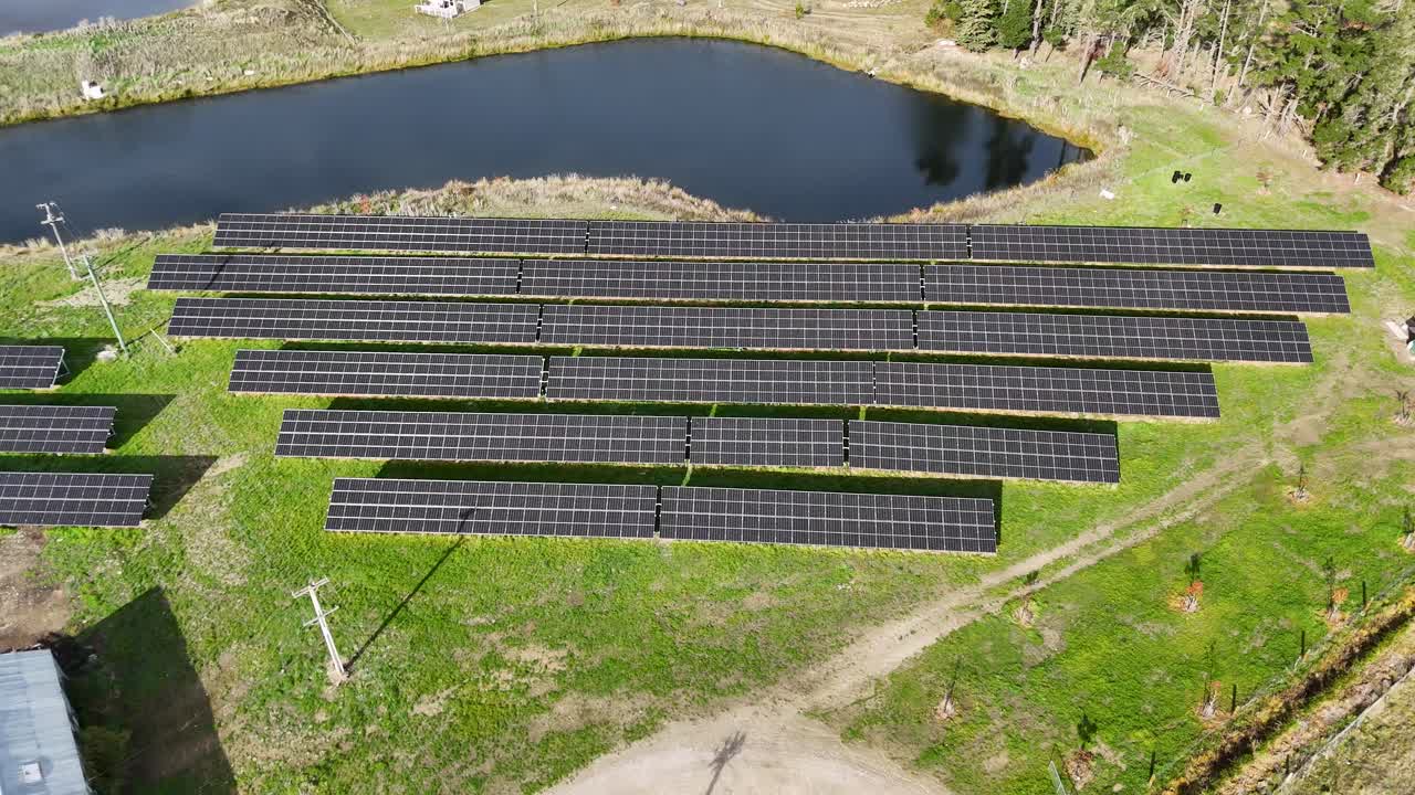 Drone footage captures a solar panel farm near a water body in Cromwell, New Zealand, under bright daylight