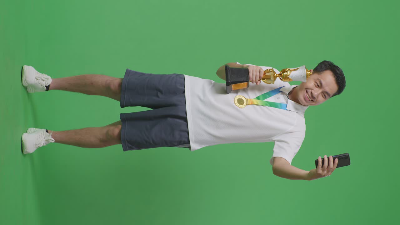 골드 트로피를 들고 있는 금메달을 가진 아시아 남자의 몸이 가득 차 있으며, 스튜디오의 녹색 스크린 배경에서 첫 번째 우승자로 스마트폰으로 사진을 찍고 있습니다.
