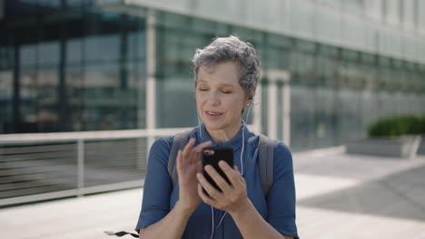 portrait-of-senior-professional-business-woman-waiting-in-city-listening-to-music-on-earphones-texting-browsing-using-smarphone-mobile-technology-in-city