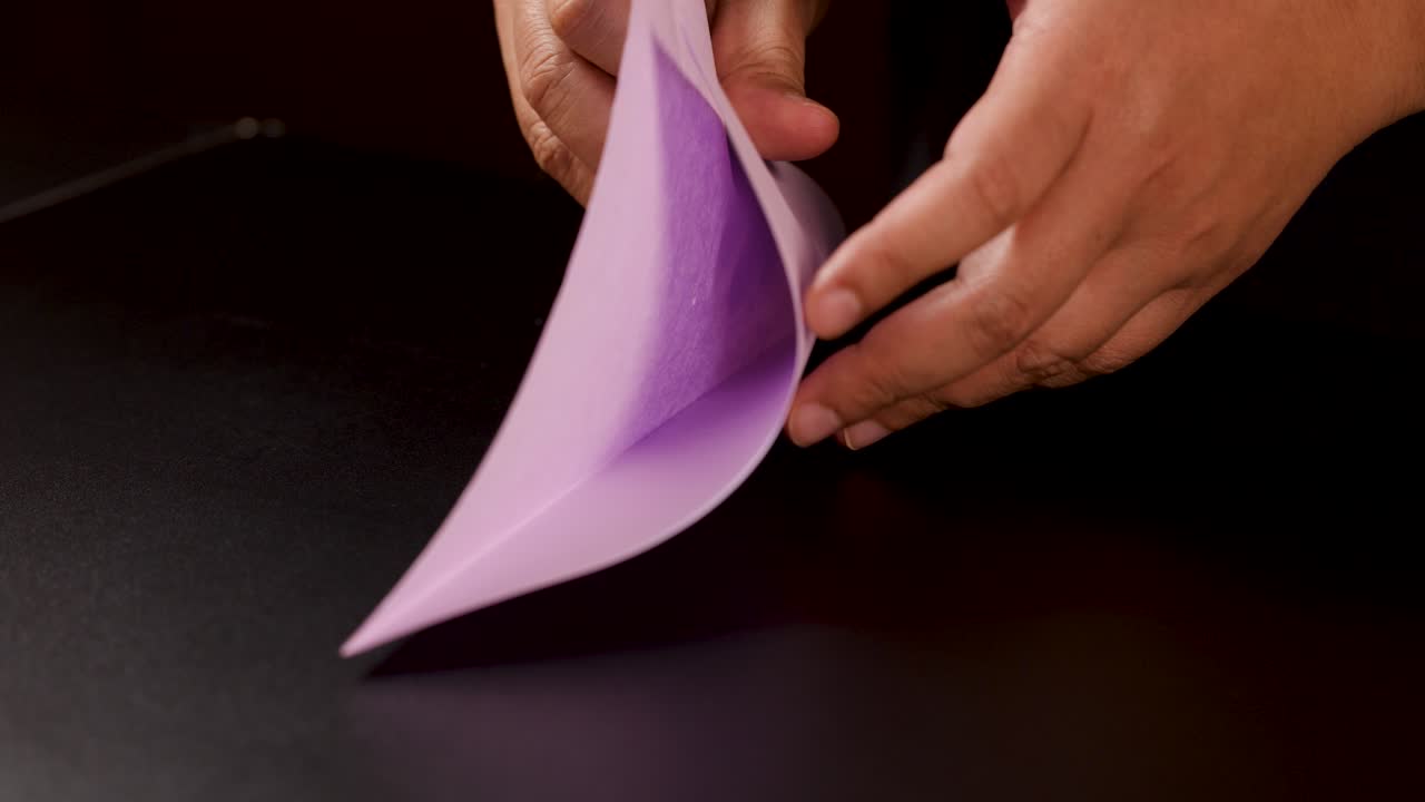 Nærbillede af hænder, der omhyggeligt folder pastelpapir til en origami-båd under blød studiebelysning