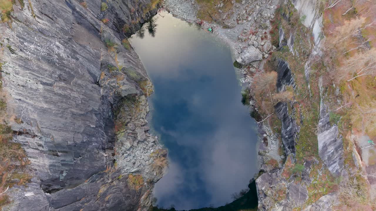 A drone looks straight down over a quarry lake as it descends, with slate walls rising around reflective water that mirrors the sky like a portal to another world