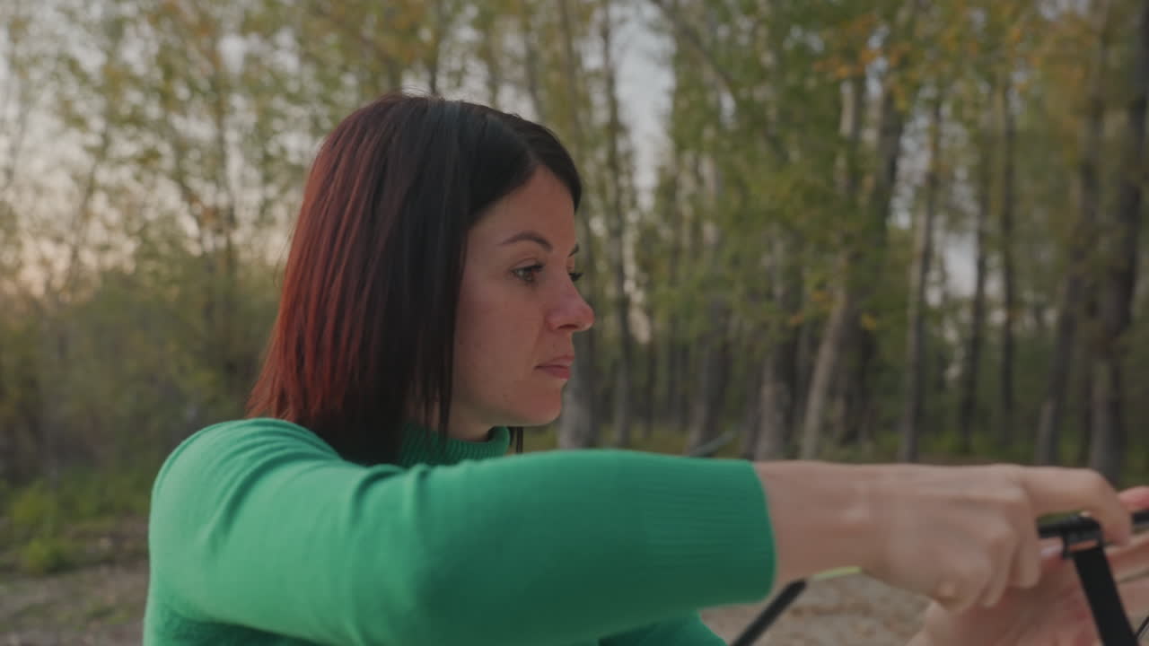 Mulher montando barraca de camping na floresta, campista morena de suéter verde alinhando postes e conectando o cubo, fundo de bétulas, luz do entardecer, ajustes cuidadosos, montagem prática ao ar livre