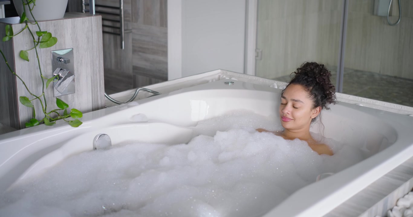 African American woman settling into foam bath in tiled bathroom, leaning back and seeking calm