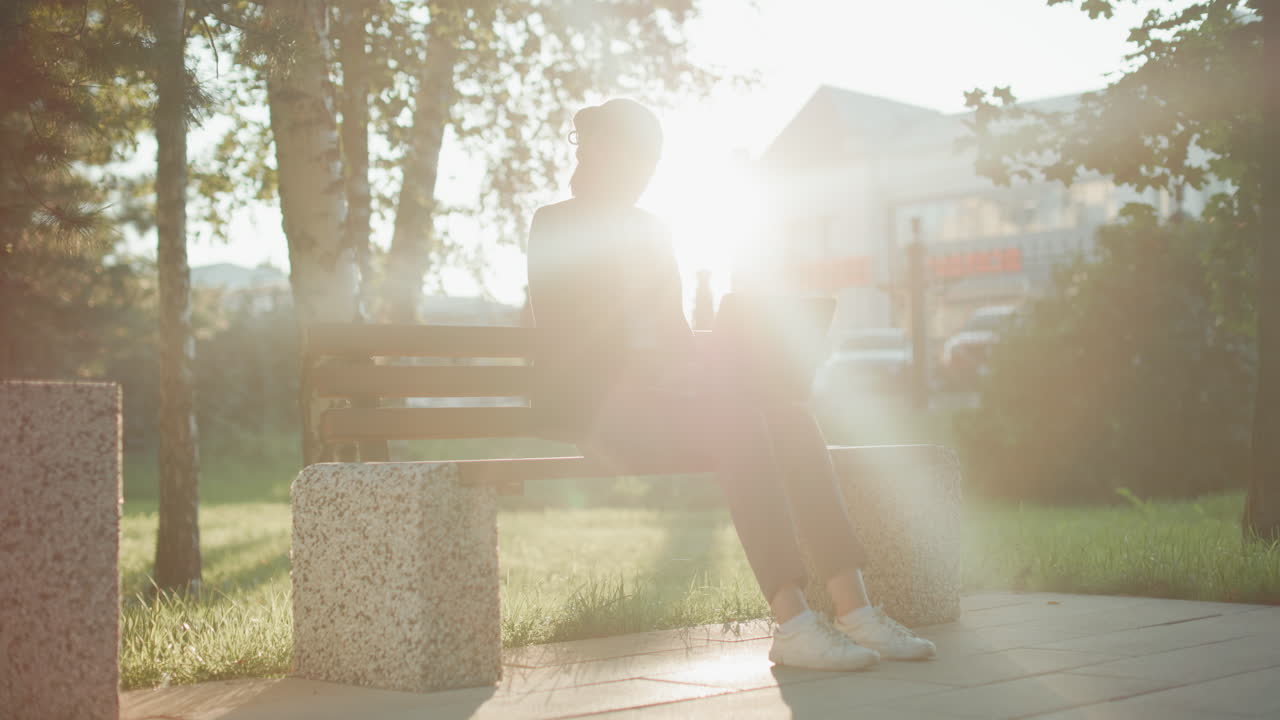 Sunlight creates glowing silhouette around woman working on laptop seated on wooden bench in outdoor park with trees and grass while buildings and parked cars are seen in distant