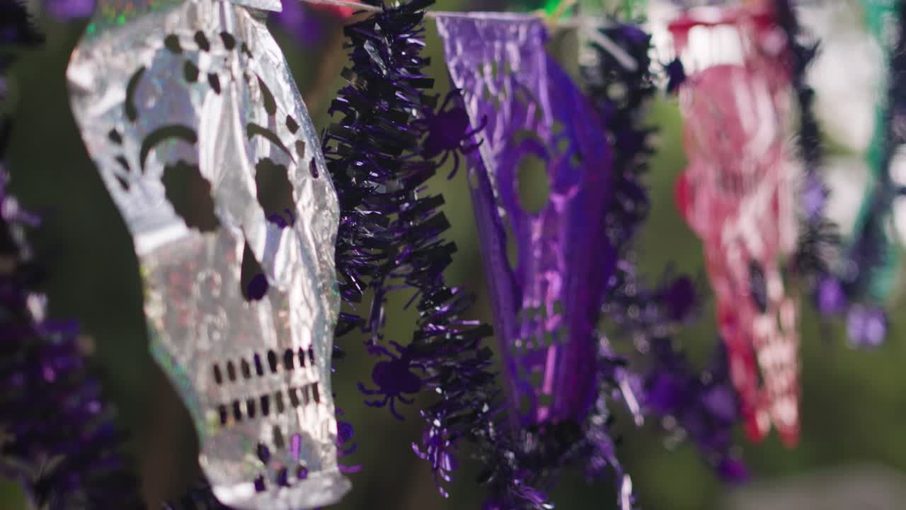 A banner of shiny skulls for Día de los Muertos hangs in cemetery with purple tinsel