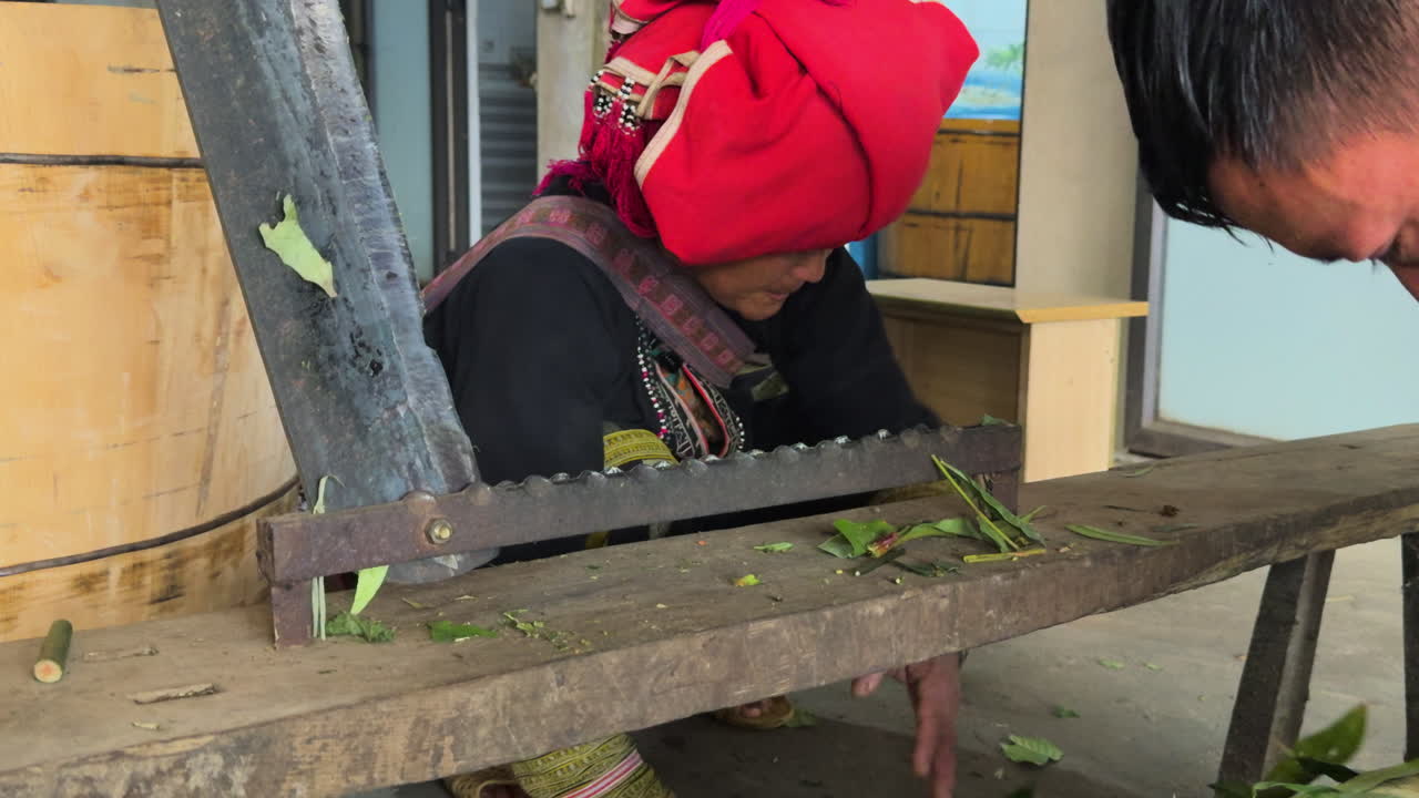 Close-up of a Black Dao man and woman working herbs through a cutting machine. Leaves and stems are separated.