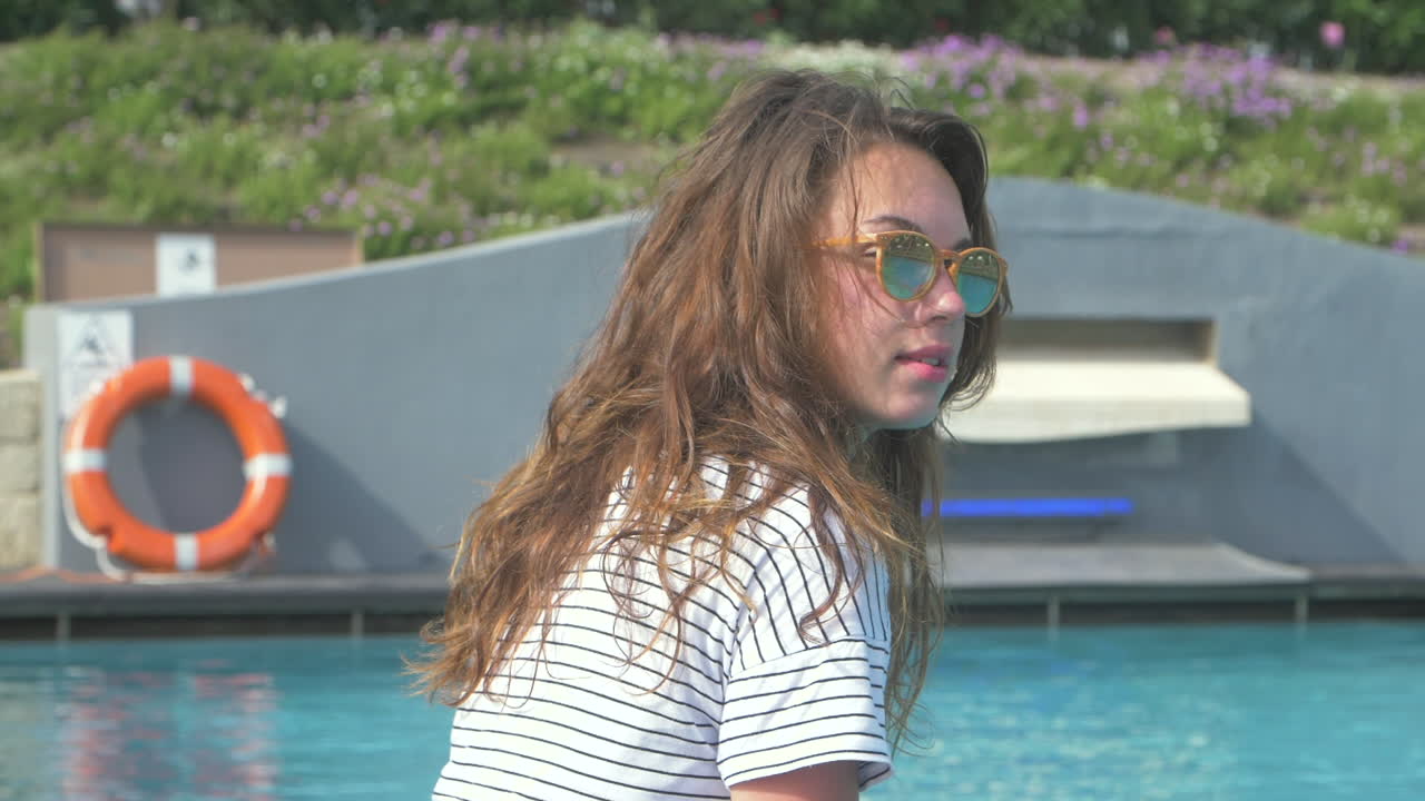 선글라스를 입은 젊은 여성이 수영장 가장자리에 앉아 웃고 있다