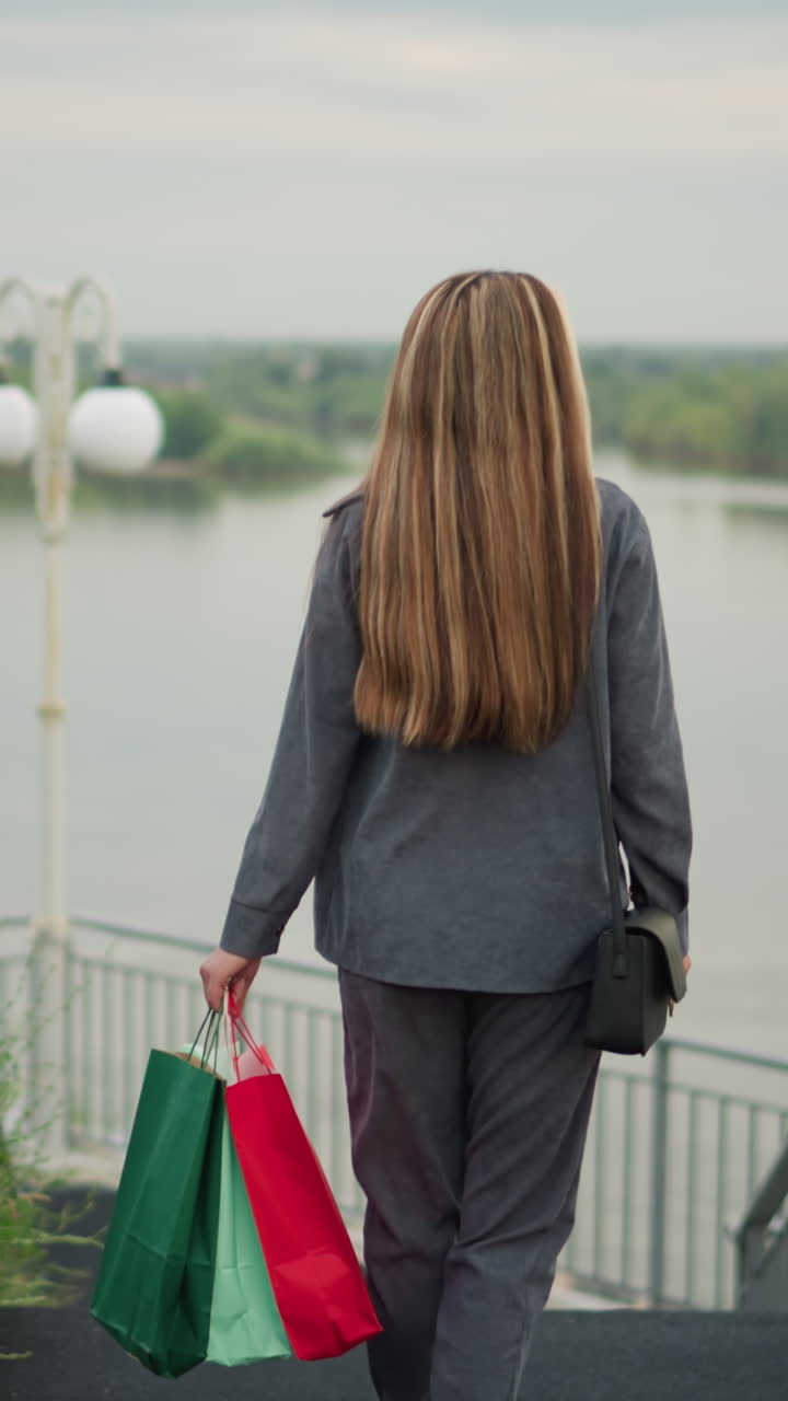 vista trasera de una mujer llevando bolsas de compras coloridas caminando por las escaleras con dos personas delante, el fondo con el río, el verde y un poste de lámpara a lo largo de las barandillas de la escalera en un día tranquilo