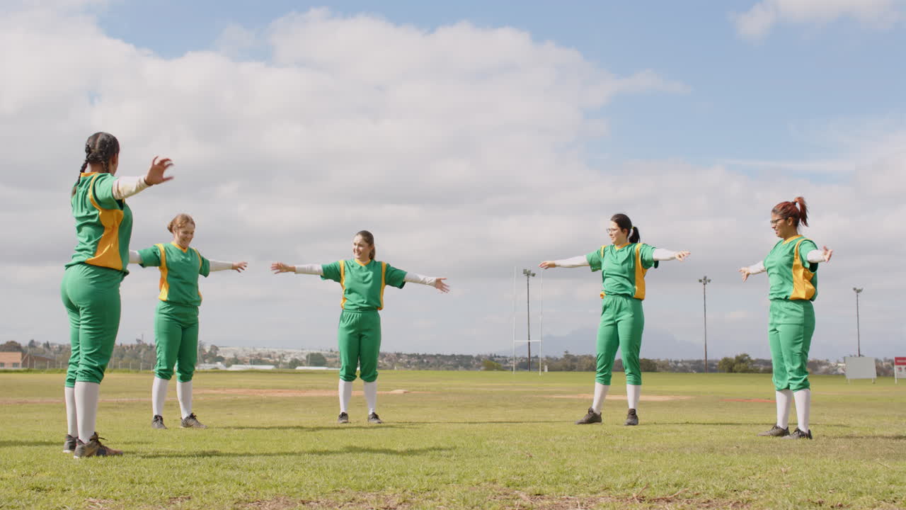 Multiracial female baseball players and male coach wearing green uniforms warming up on a pitch