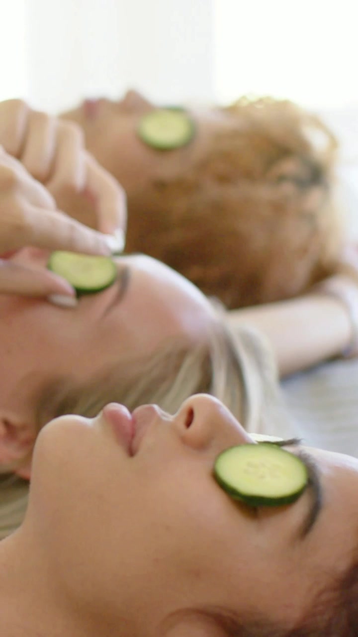Vertikal video: En hånd placerer agurkeskiver på tre kvinder, der ligger i badekåber på spa til afslapning.