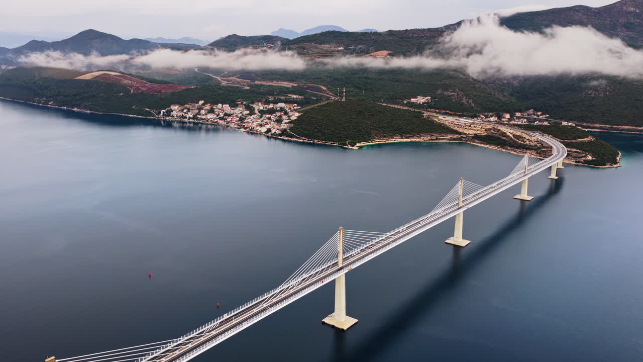 Aerial drone view of the Peljesac Bridge near Komarna, Croatia, partially shrouded in low coastal fog with mountains in the background