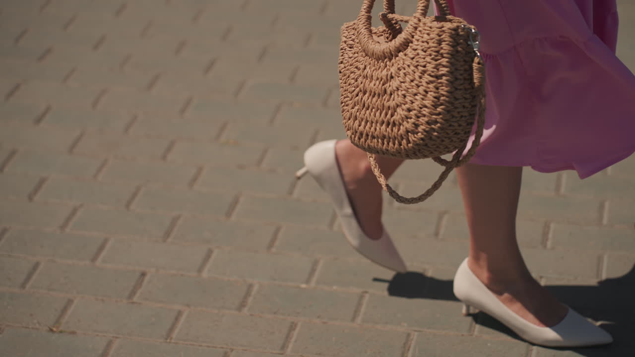 Primo piano di una donna che cammina indossando un abito rosa, una borsa di paglia intrecciata e scarpe basse bianche su un marciapiede soleggiato. Atmosfera da shopper rilassata con dettagli di moda slow, manico testurizzato della tote bag, passo leggero che proietta un'ombra lunga, spontanea