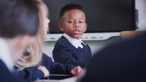 Colegial-Negro-Sentado-En-La-Mesa-Hablando-Con-Compañeros-De-Clase-En-Una-Lección-De-Escuela-Primaria,-Enfoque-Selectivo