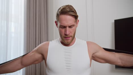 straight view of a blond athletic man with stubble a man in a white t-shirt does morning exercises and swings his arms using a special rubber expander at home in a modern bright apartment at home