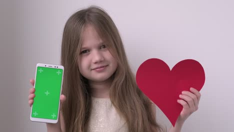 cute child girl shows phone with green screen mock up cellphone, mobile, telephone and red heart. chroma key green screen smartphone set up for advertising. valentines day, love, mothers day