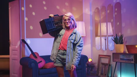 happy cheerful young caucasian woman in glasses standing in living room in a retro party at home holding retro boom box on a shoulder and looking at camera