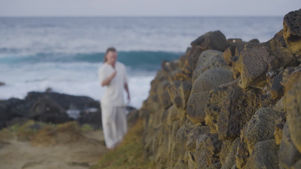 A sharp focus on a textured stone wall contrasts with a blurred man in white standing near the ocean horizon, waves breaking softly behind him