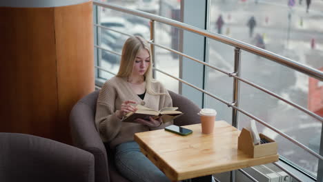 lady seated at table, thoughtfully reading book and flipping to next page, with coffee cup, phone, and napkin on table, modern urban cafe setting with large window view outside