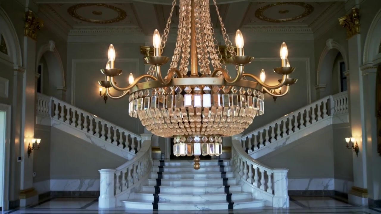 Telecamera che si allontana, rivelando un lampadario a più livelli ornato in un grande ingresso, per mostrare una scala in marmo.