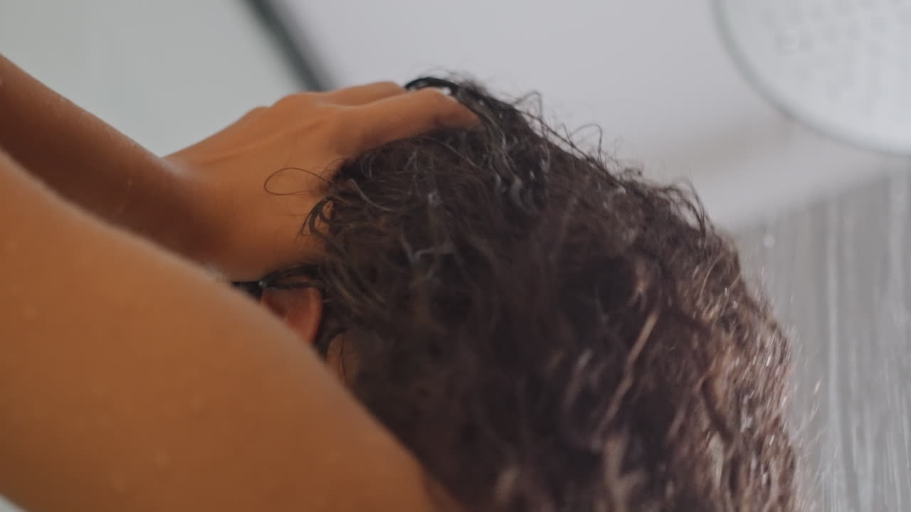 Donna nella doccia dell'hotel che lava i capelli ricci, delicato massaggio al cuoio capelluto sotto il soffione a pioggia, goccioline calde sulle spalle, articoli da toeletta color ambra sullo scaffale, luce diurna soffusa, relax nel bagno di un hotel boutique.