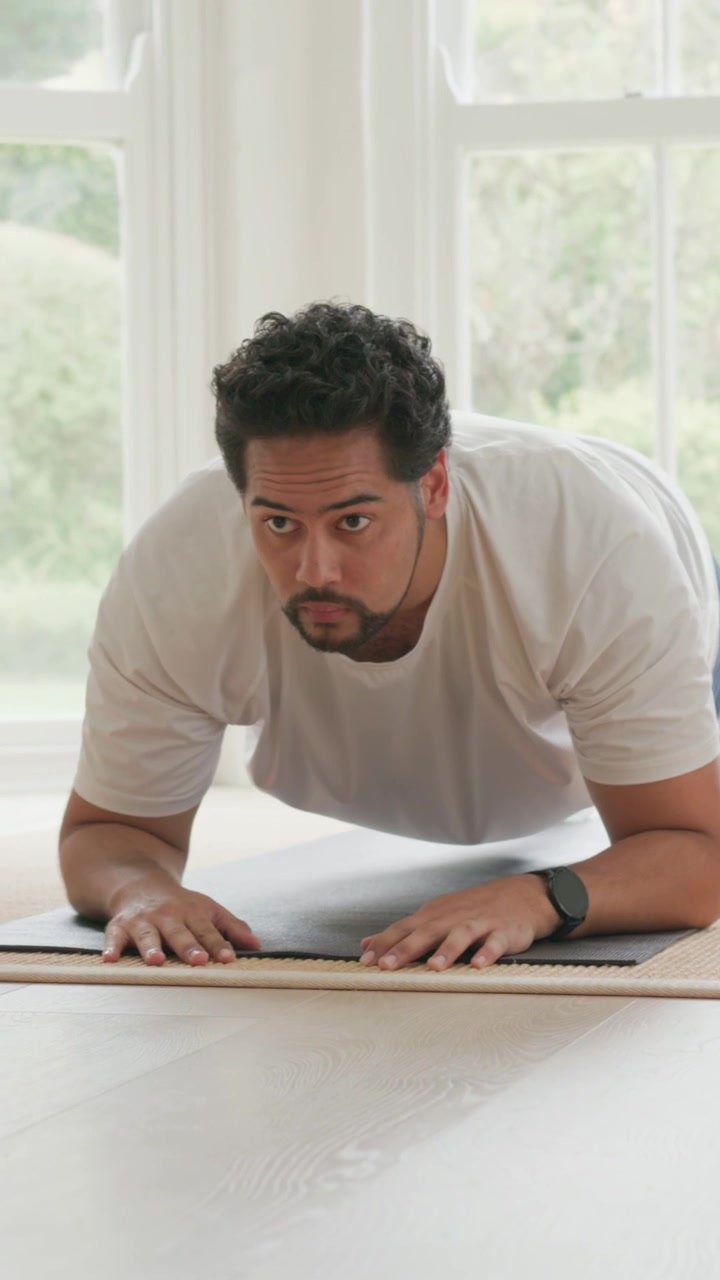 Vertical video: Entering front room man in shorts approaching mat holding forearm plank for fitness