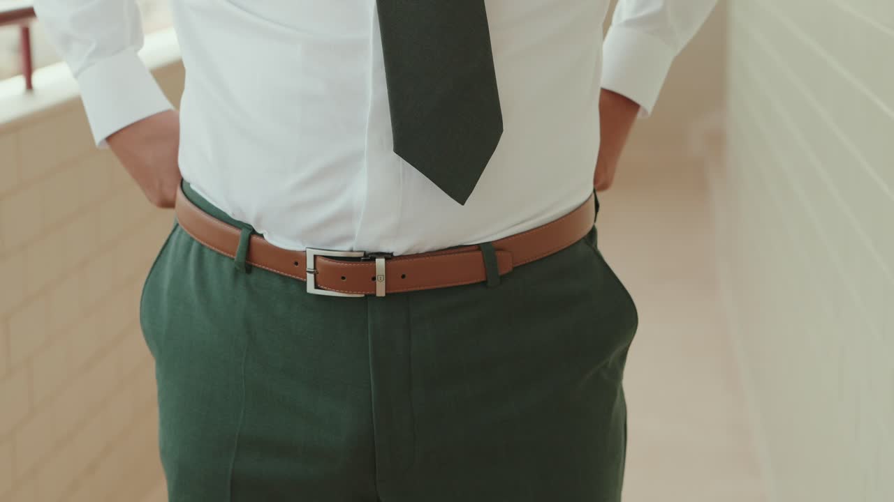 Close up of groom adjusting brown belt on green trousers while wearing white shirt and dark tie