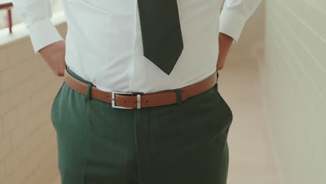 close up of groom adjusting brown belt on green trousers while wearing white shirt and dark tie