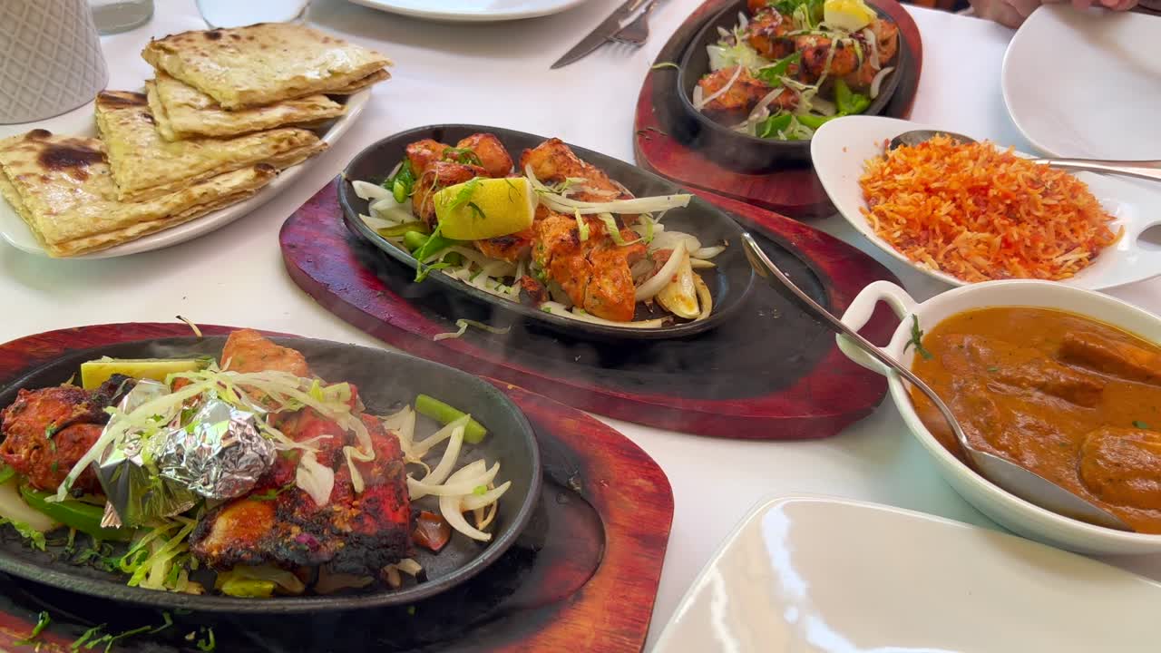Traditional Indian Tandoori chicken with onions on a very hot plate with steam smoke, butter chicken, cheese naan bread and coconut rice in a restaurant, authentic asian food, 4K shot