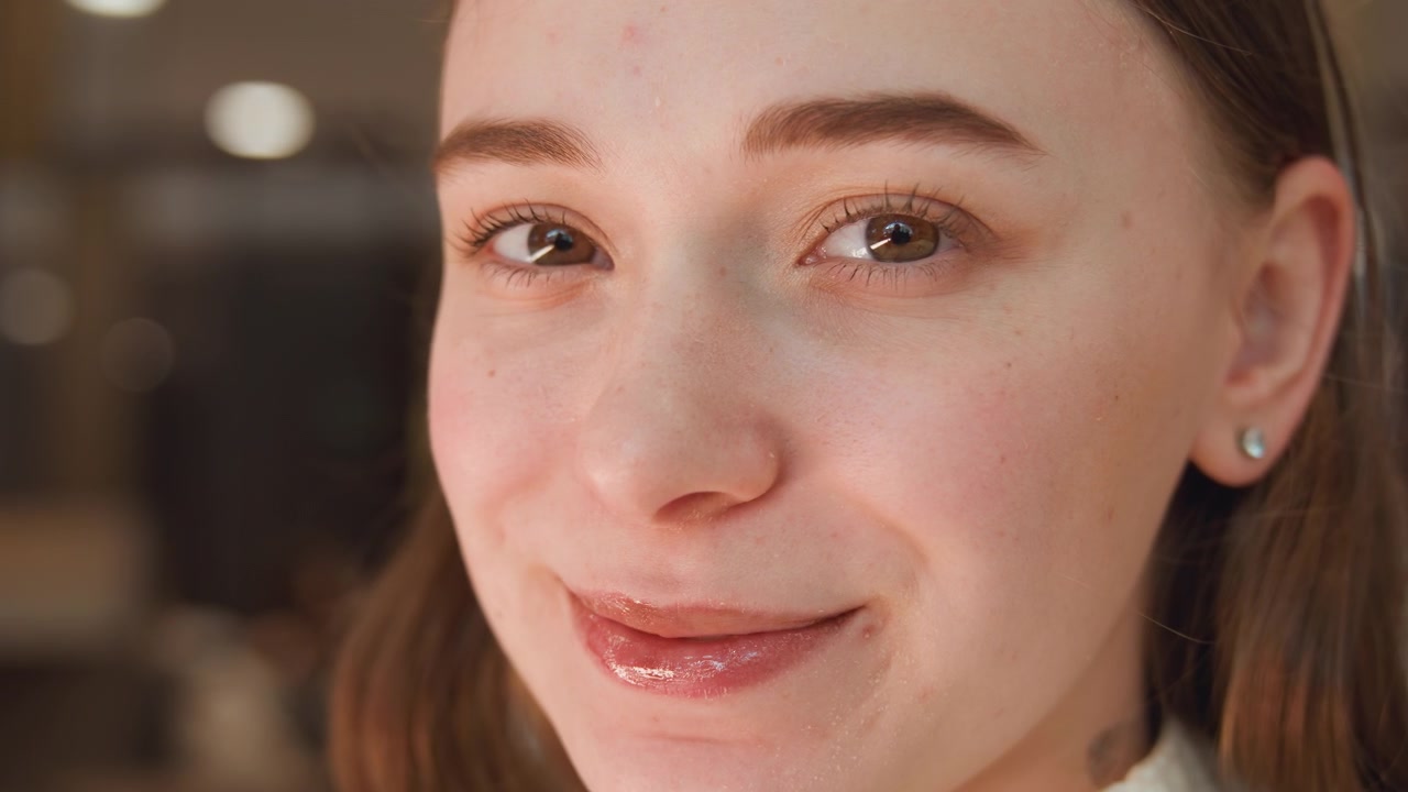 Close head and shoulders view of young woman applying liquid lip color with wand applicator, showing focused hand movement, soft indoor light, retail vanity background and relaxed beauty routine