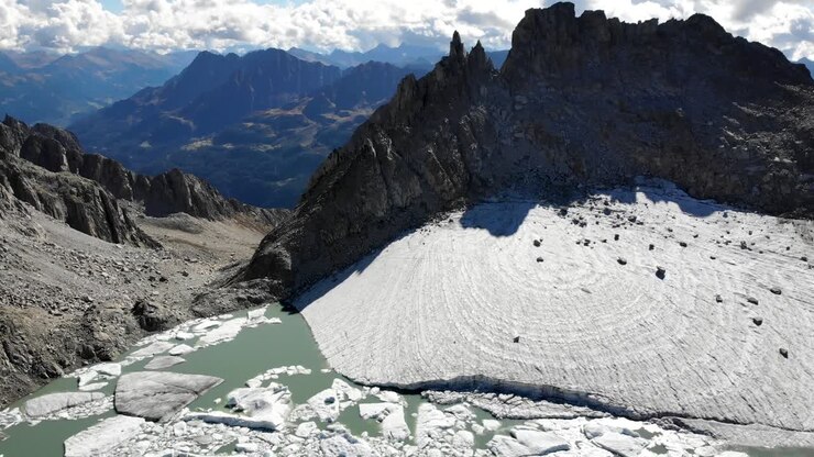 Aerial flyover above a glacier lake full of melted icebergs in remote parts of the Swiss Alps on a sunny day with the view panning down from the mountain peaks to the lake shore