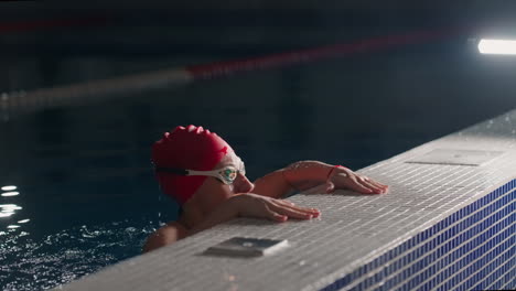 a female swimmer in a red cap and goggles is resting on the edge of a pool