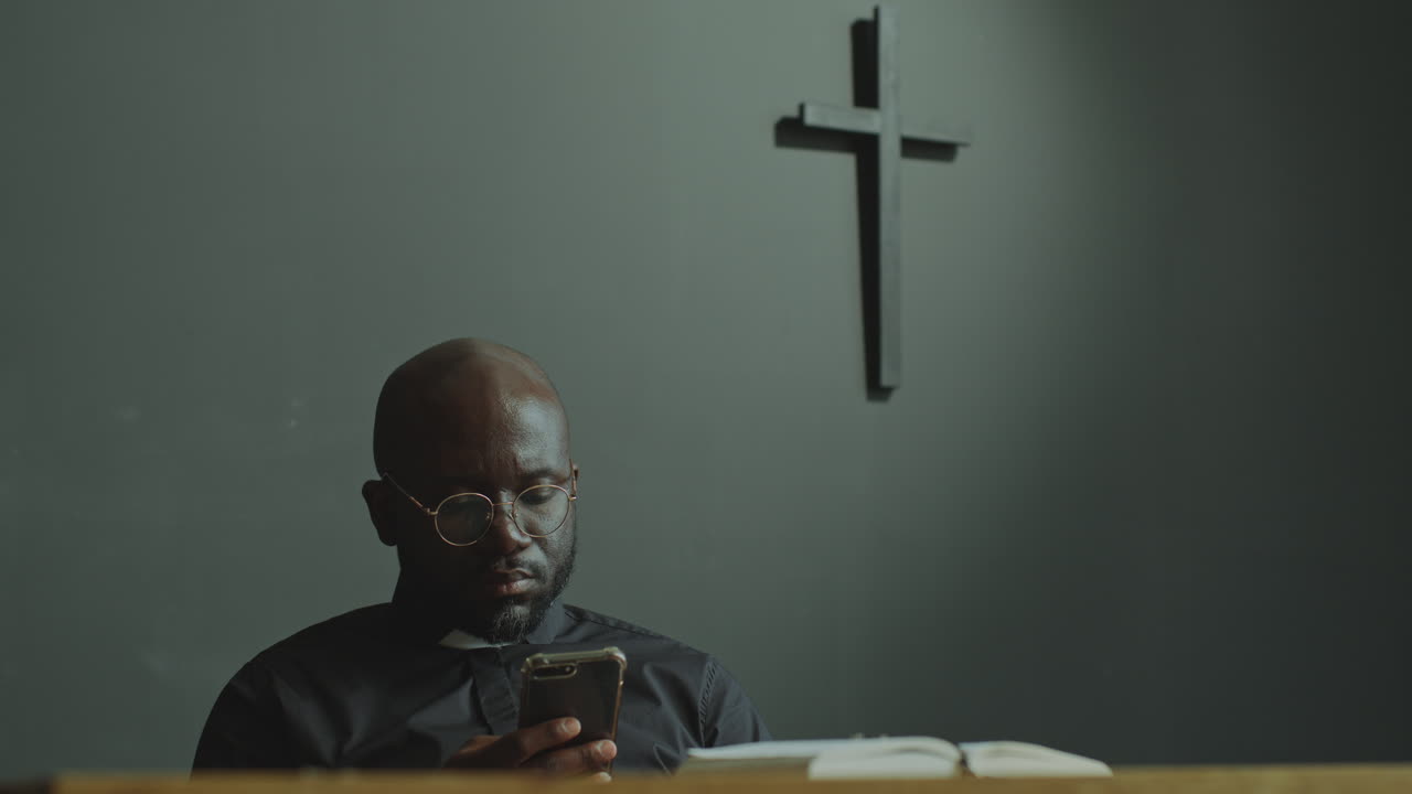 Černý katolický kněz používající mobilní telefon v moderním kostele