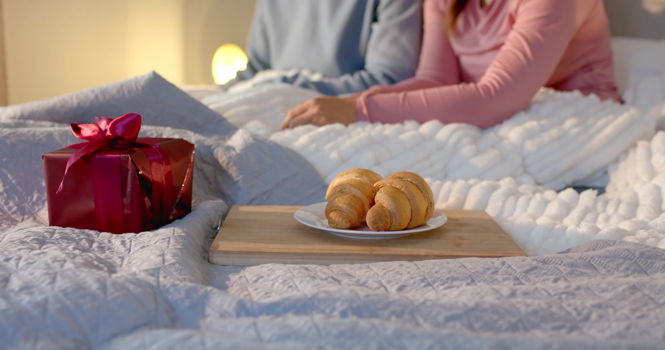 Couple smoothing blankets on bed after spotting breakfast tray holding croissants and gift box