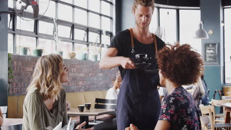 two female friends sitting at table in coffee shop being served by waiter