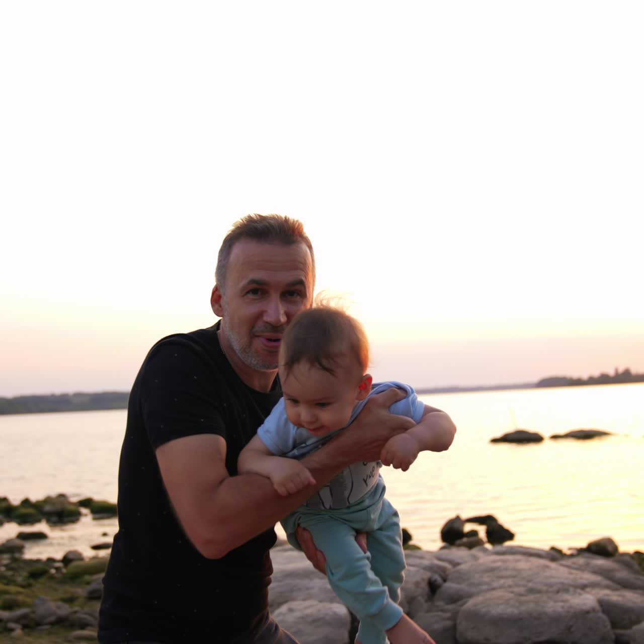 Father playing with little son at the shore. Cute baby boy is waved in dad's hands closer to camera. Clear sky at backdrop
