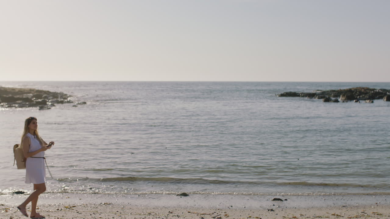 은 해변에서 걸어다니는 젊은 여성이 휴가를 즐기고 있는 사진을 찍고 있습니다.