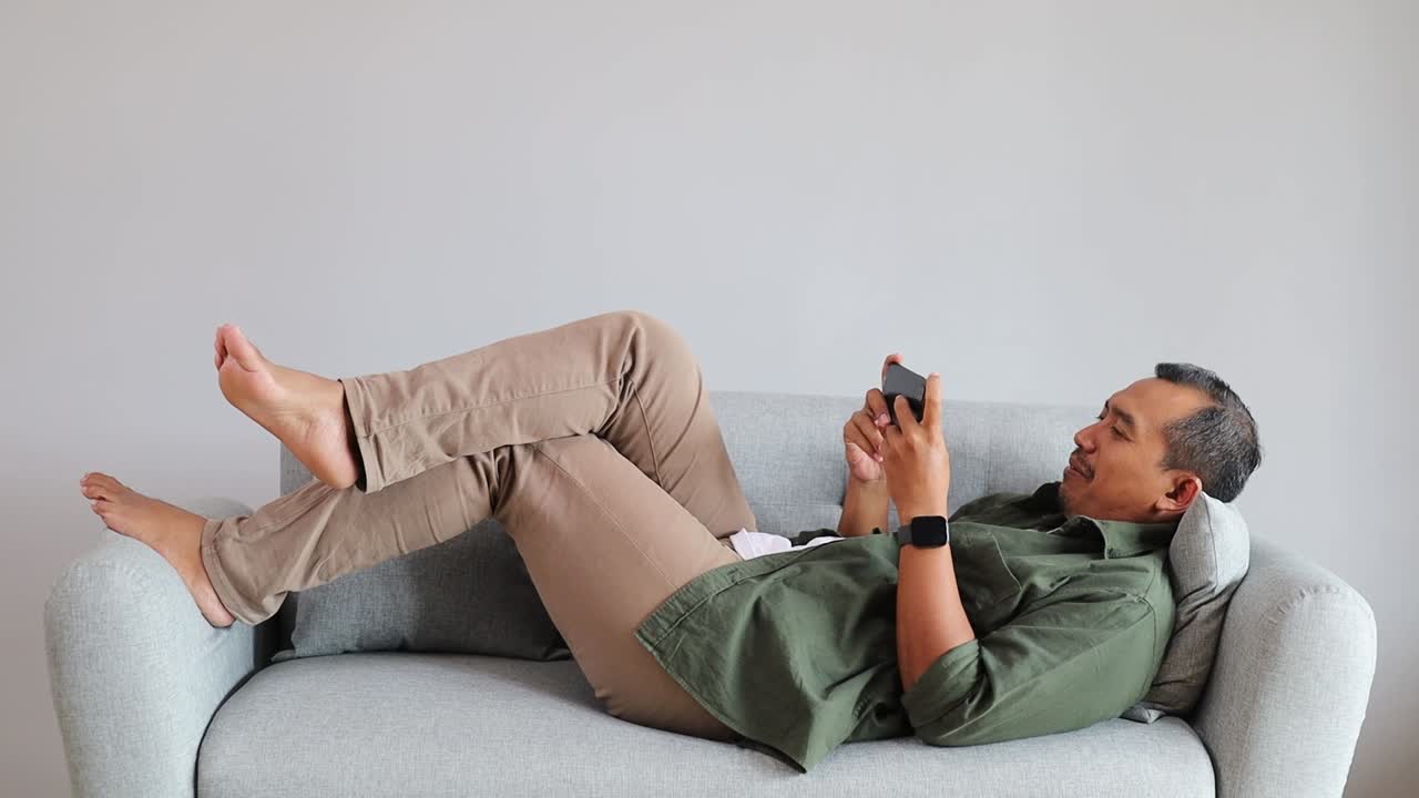 Uomo asiatico felice che gioca online al telefono cellulare sdraiato sul divano di casa.
