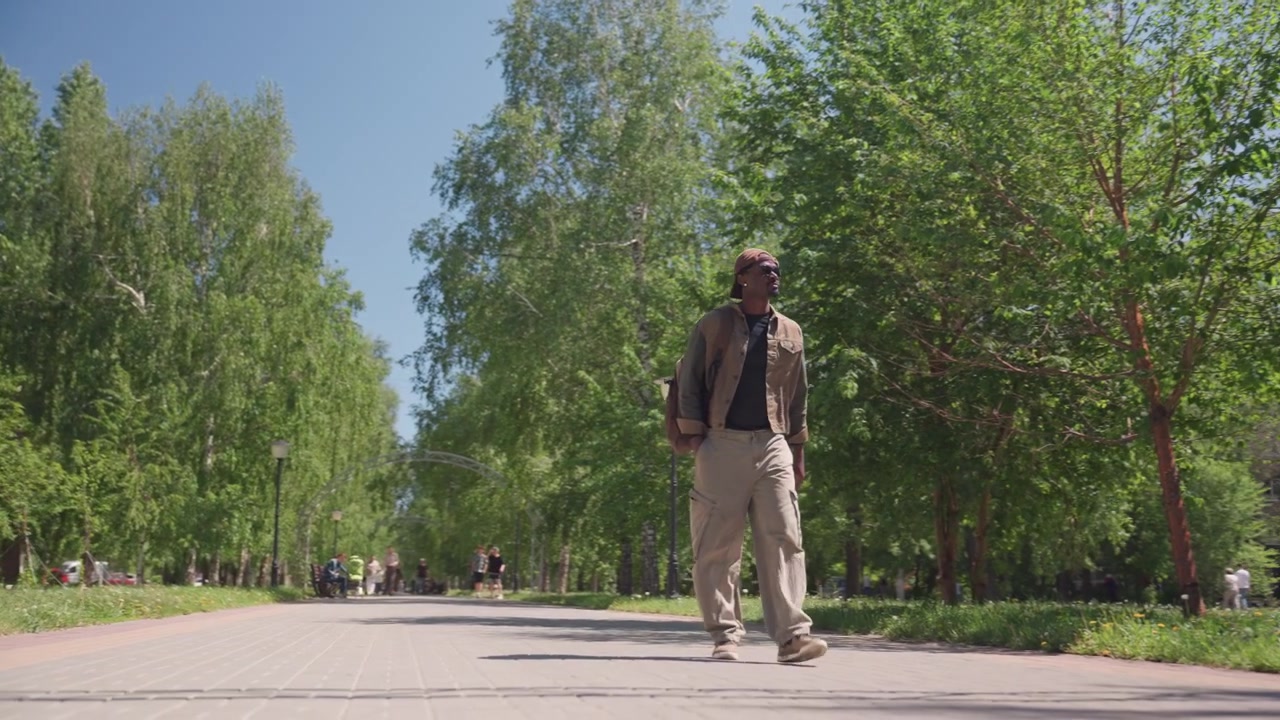 Man walks steadily along paved path in urban park wearing casual outdoor clothing and carrying bag. scene features green trees lining pathway with parked cars visible near residential