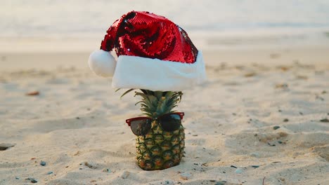 Strand-Auf-Curaçao-–-Ananas-Mit-Roter-Weihnachtsmütze-Und-Sonnenbrille-Am-Sandstrand-Mit-Wellen-Im-Hintergrund-–-Nahaufnahme