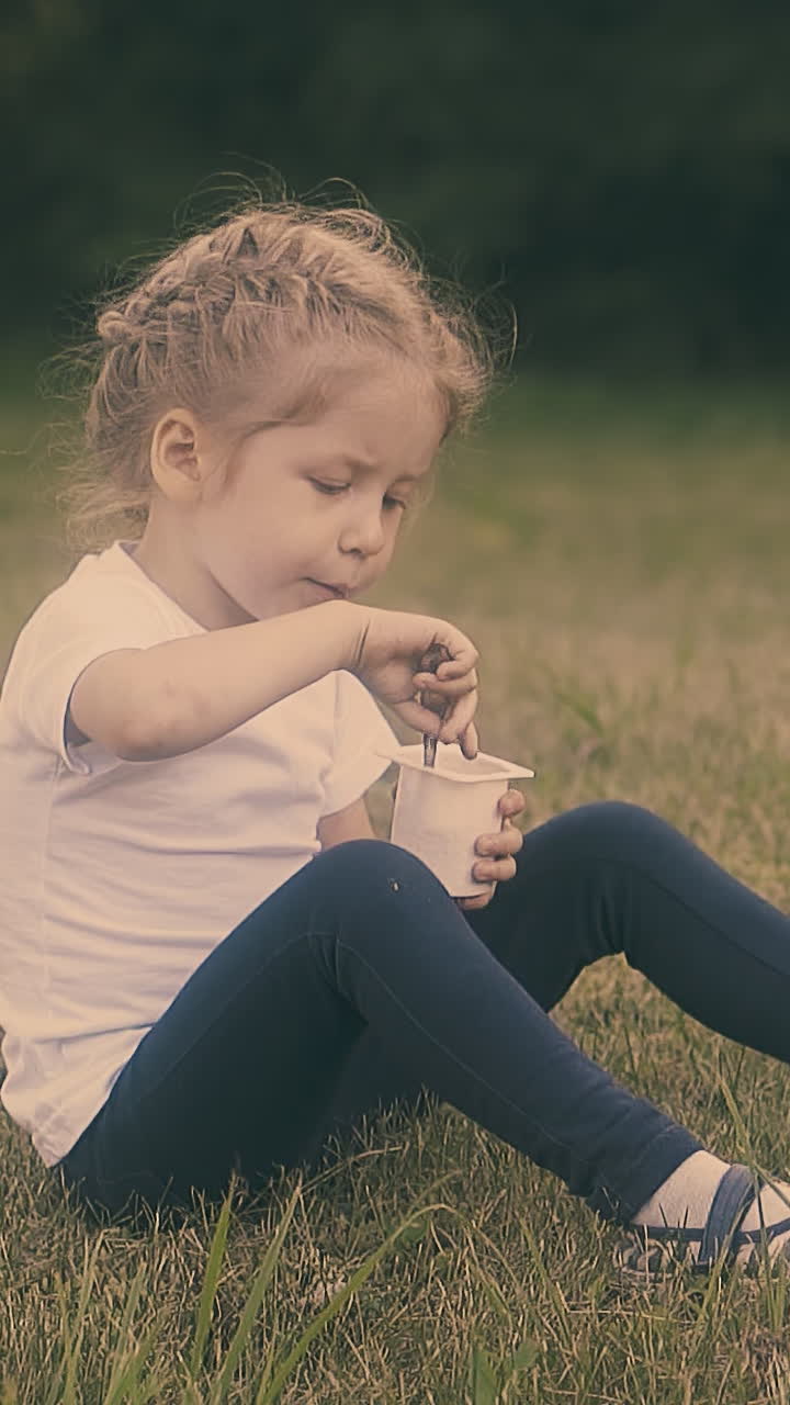 cute blond little girl eats fresh yogurt with spoon resting on grass lawn in green sunny park closeup slow motion