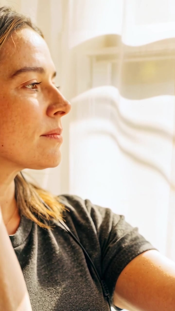 Vertical video: Noticing sun, woman in grey tee with headphones pulling curtain at window, gazing